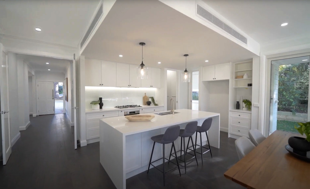 This is an example of a large l-shaped open plan kitchen in Melbourne with a submerged sink, shaker cabinets, white cabinets, white splashback, glass sheet splashback, stainless steel appliances, dark hardwood flooring, an island and a drop ceiling.