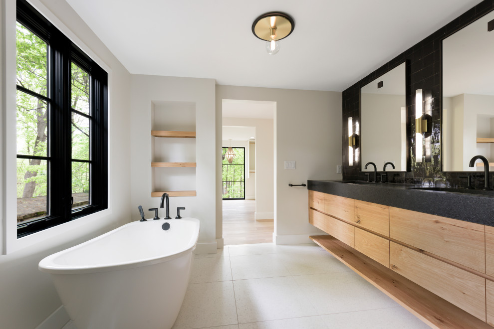 Photo of a large midcentury master bathroom in Minneapolis with medium wood cabinets, flat-panel cabinets, a freestanding tub, an alcove shower, a one-piece toilet, black tile, cement tile, white walls, terrazzo floors, an undermount sink, granite benchtops, white floor, a hinged shower door and black benchtops.
