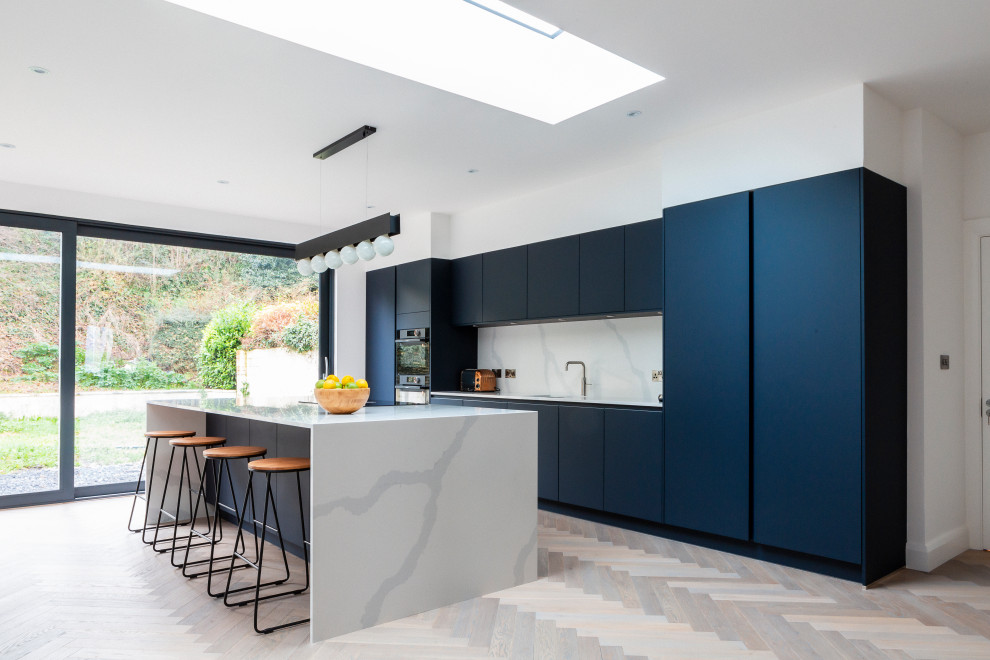 This is an example of a large contemporary galley kitchen in Dublin with a single-bowl sink, flat-panel cabinets, stone slab splashback, black appliances, light hardwood floors, blue cabinets, white splashback, with island, beige floor and white benchtop.