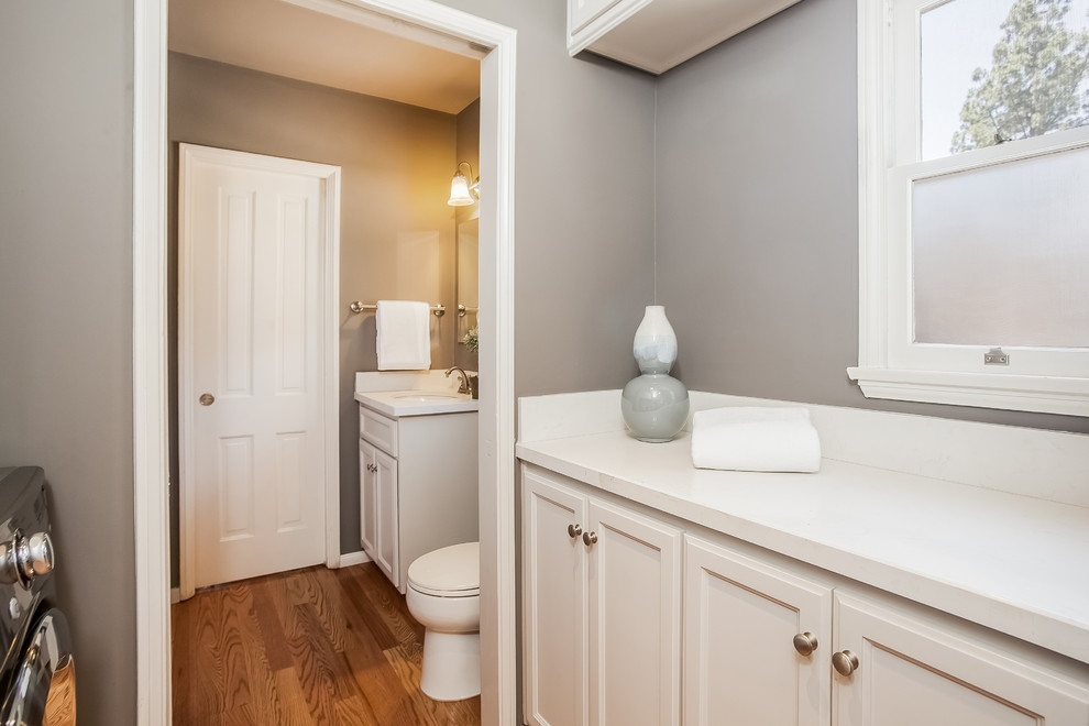 Large transitional l-shaped laundry room in Los Angeles with an undermount sink, shaker cabinets, white cabinets, light hardwood floors, quartzite benchtops, grey walls and a side-by-side washer and dryer.
