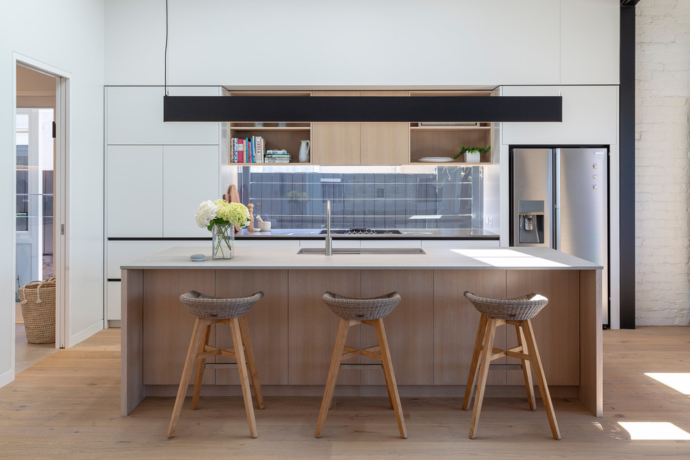 Mid-sized contemporary single-wall open plan kitchen in Auckland with an integrated sink, white cabinets, quartz benchtops, stainless steel appliances, light hardwood floors, with island, brown floor, grey benchtop, flat-panel cabinets and window splashback.