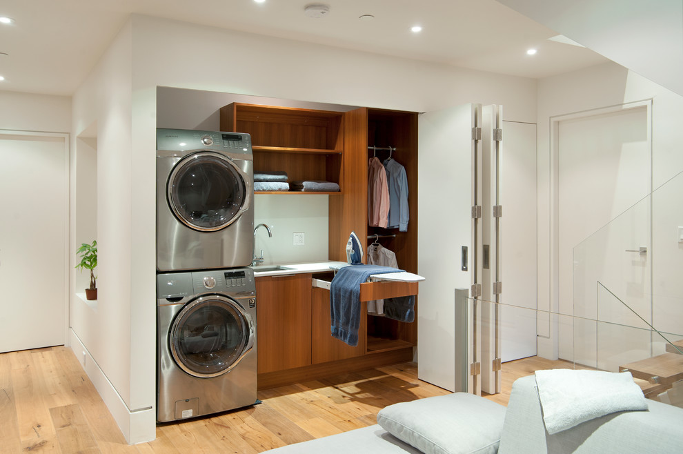 Design ideas for a contemporary single-wall laundry cupboard in Vancouver with an undermount sink, flat-panel cabinets, medium wood cabinets, white walls, medium hardwood floors, a stacked washer and dryer and white benchtop.