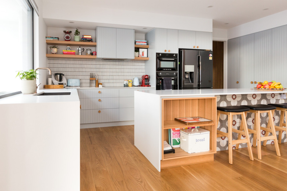 Photo of a midcentury l-shaped kitchen in Canberra - Queanbeyan with an undermount sink, flat-panel cabinets, grey cabinets, white splashback, mosaic tile splashback, medium hardwood floors, with island, brown floor and white benchtop.