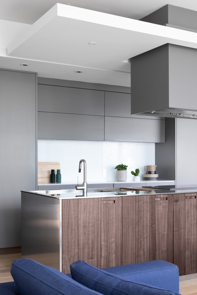 This is an example of a mid-sized eclectic single-wall open plan kitchen in Other with an integrated sink, flat-panel cabinets, grey cabinets, stainless steel benchtops, white splashback, panelled appliances, light hardwood floors, with island and beige floor.