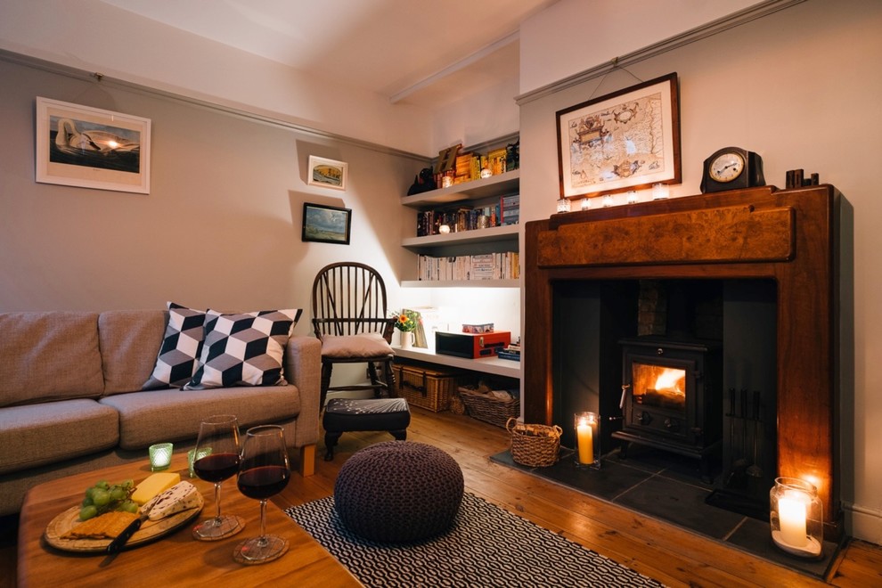 Photo of a mid-sized country enclosed living room in Cornwall with a library, beige walls, light hardwood floors, a wood stove and a wood fireplace surround.