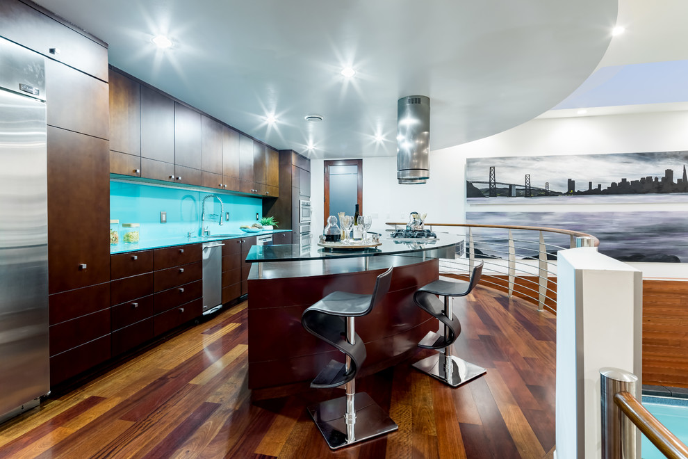 Photo of a large contemporary open plan kitchen in San Francisco with a drop-in sink, flat-panel cabinets, brown cabinets, glass benchtops, black splashback, glass sheet splashback, stainless steel appliances, with island, brown floor, blue benchtop and medium hardwood floors.