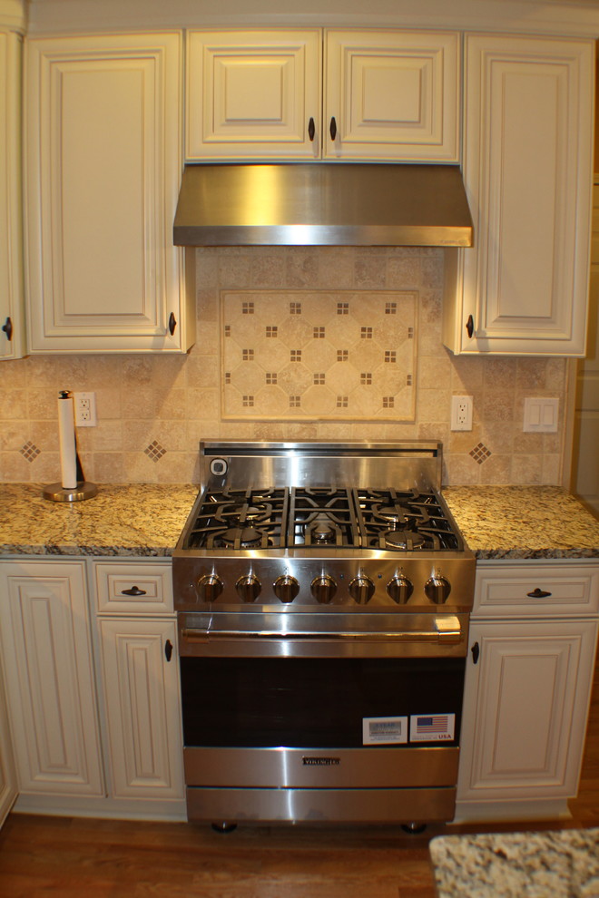 Example of a transitional medium tone wood floor kitchen design with raised-panel cabinets, white cabinets, granite countertops, beige backsplash, stone tile backsplash, stainless steel appliances and an island