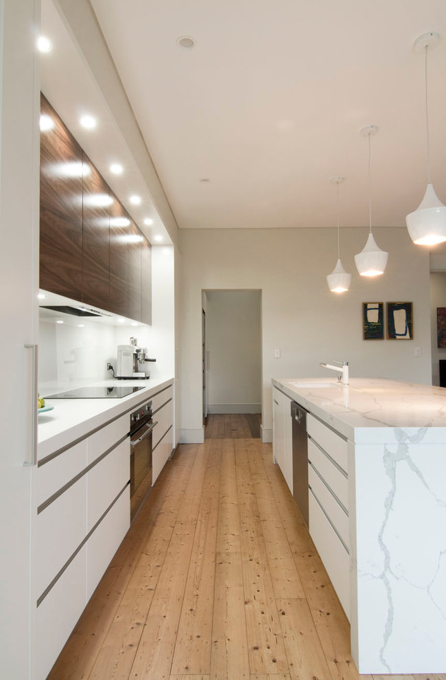 This is an example of a large modern single-wall open plan kitchen in Sydney with an undermount sink, flat-panel cabinets, quartz benchtops, white splashback, glass sheet splashback, stainless steel appliances, light hardwood floors, with island and medium wood cabinets.