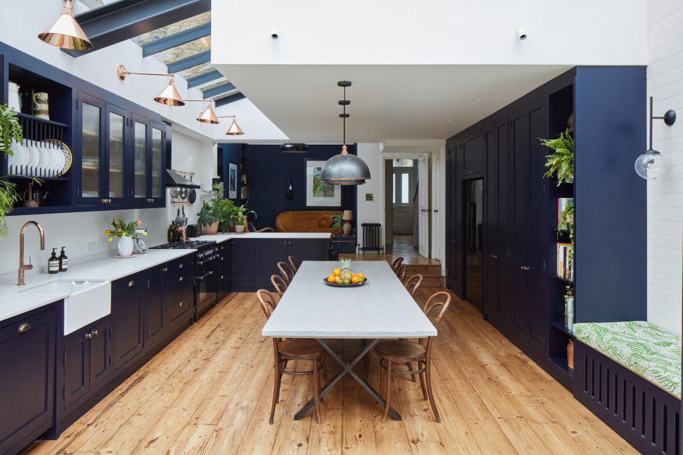 This is an example of a large transitional l-shaped eat-in kitchen in London with a farmhouse sink, shaker cabinets, blue cabinets, quartz benchtops, white splashback, engineered quartz splashback, black appliances, light hardwood floors, no island, grey benchtop, exposed beam and brown floor.