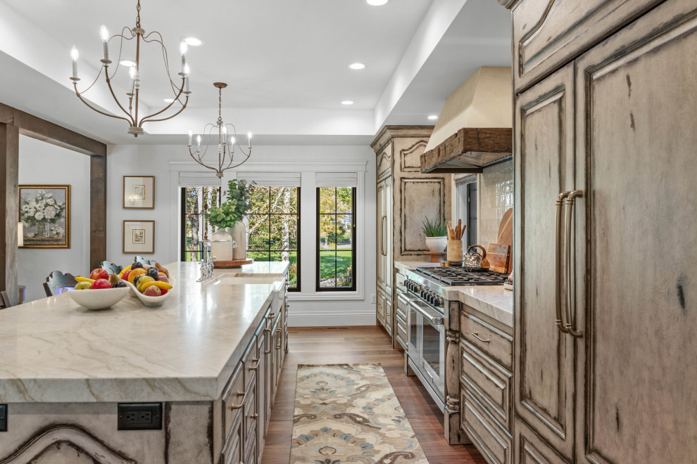 Inspiration for a large single-wall open plan kitchen in Other with a farmhouse sink, raised-panel cabinets, distressed cabinets, quartzite benchtops, white splashback, ceramic splashback, panelled appliances, medium hardwood floors, with island, brown floor, beige benchtop and recessed.