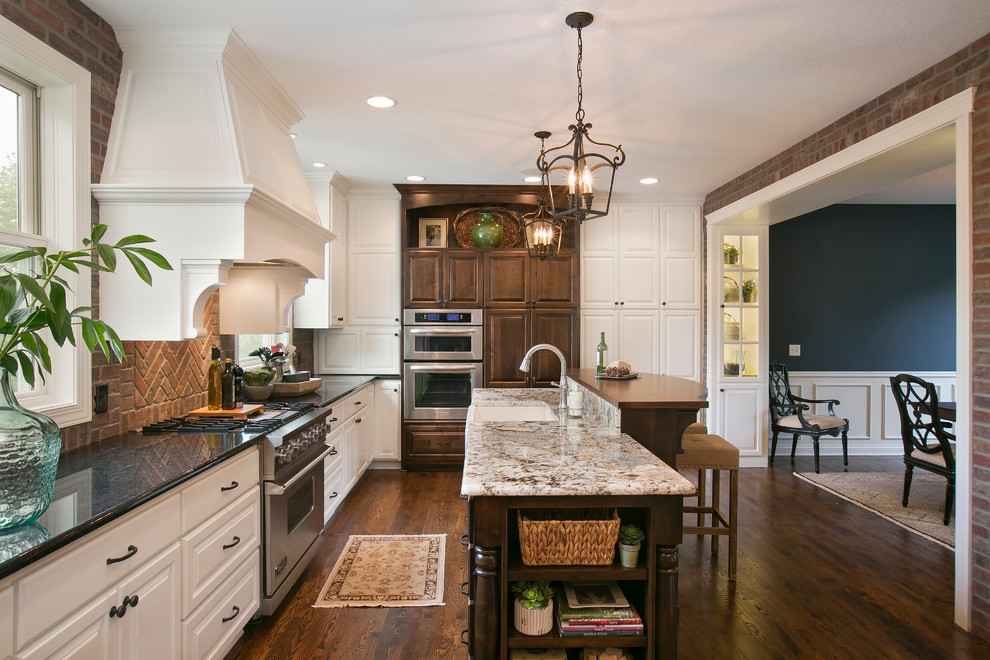 Photo of a mid-sized traditional u-shaped eat-in kitchen in Minneapolis with a farmhouse sink, raised-panel cabinets, white cabinets, granite benchtops, red splashback, brick splashback, stainless steel appliances, dark hardwood floors, with island, brown floor and multi-coloured benchtop.