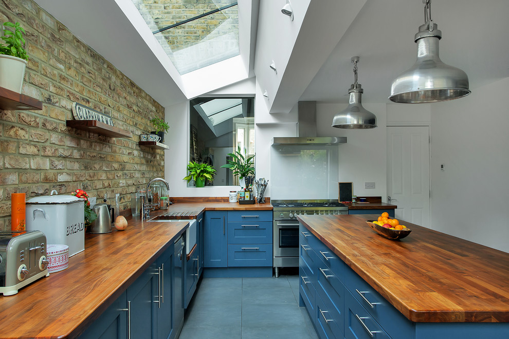 Large industrial eat-in kitchen in London with a farmhouse sink, shaker cabinets, blue cabinets, wood benchtops, stainless steel appliances, ceramic floors, with island, grey floor and brown benchtop.