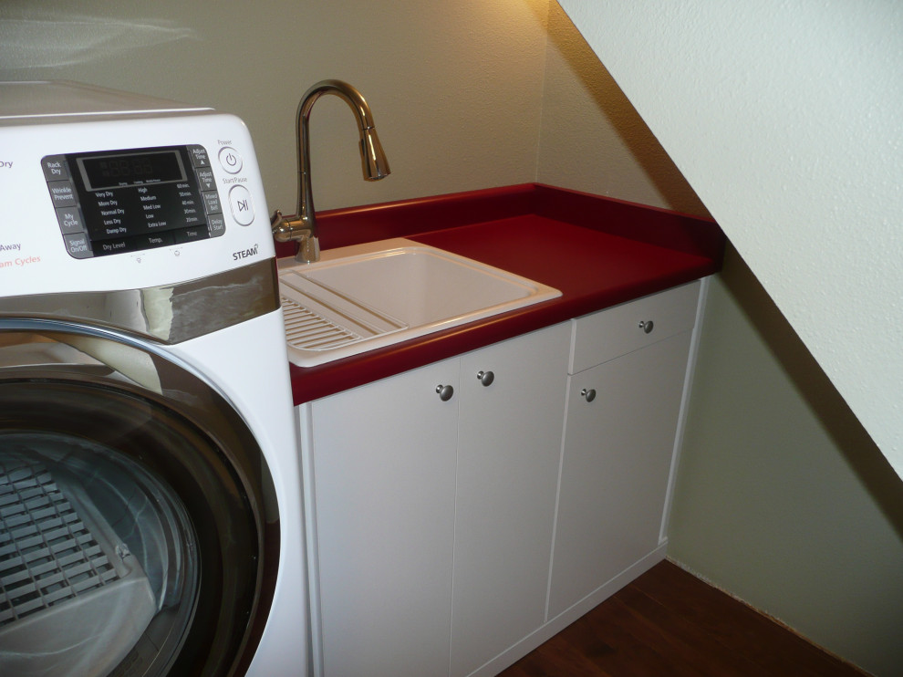 シアトルにあるお手頃価格の小さなトランジショナルスタイルのおしゃれな家事室 (コの字型、ドロップインシンク、フラットパネル扉のキャビネット、白いキャビネット、ラミネートカウンター、赤いキッチンパネル、緑の壁、無垢フローリング、左右配置の洗濯機・乾燥機、茶色い床、赤いキッチンカウンター) の写真