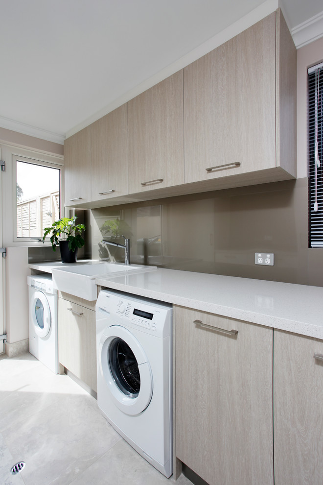 Photo of a modern single-wall laundry room in Perth with a farmhouse sink, flat-panel cabinets, quartz benchtops, light wood cabinets, white benchtop, a side-by-side washer and dryer, ceramic floors and grey floor.