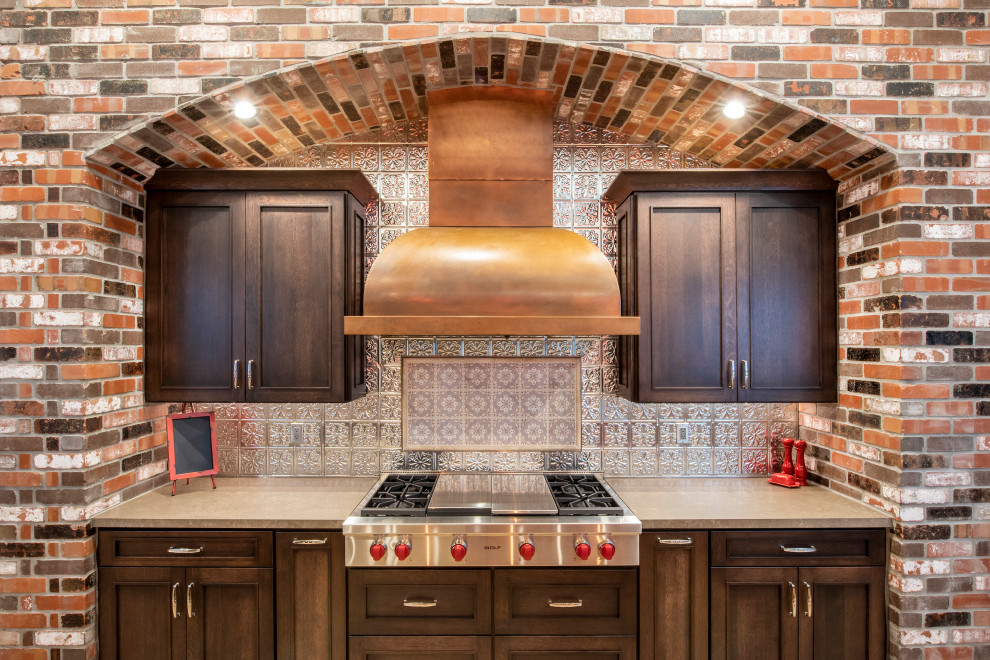 Photo of an expansive arts and crafts l-shaped open plan kitchen in Phoenix with a farmhouse sink, shaker cabinets, brown cabinets, granite benchtops, metallic splashback, metal splashback, stainless steel appliances, travertine floors, multiple islands, beige floor and red benchtop.