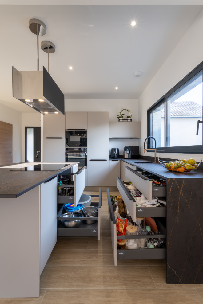 This is an example of a mid-sized contemporary l-shaped open plan kitchen in Lyon with an integrated sink, beaded inset cabinets, beige cabinets, tile benchtops, black splashback, ceramic splashback, panelled appliances, light hardwood floors, with island, brown floor and black benchtop.