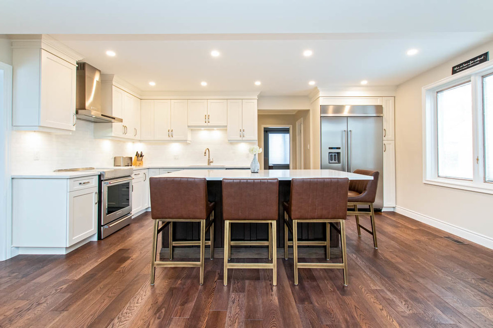 Example of a mid-sized transitional l-shaped medium tone wood floor and brown floor open concept kitchen design with an undermount sink, shaker cabinets, white cabinets, quartzite countertops, white backsplash, stone tile backsplash, stainless steel appliances, an island and white countertops