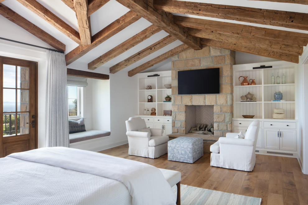 Large country master bedroom in Santa Barbara with white walls, medium hardwood floors, a standard fireplace, a stone fireplace surround, brown floor, exposed beam and vaulted.