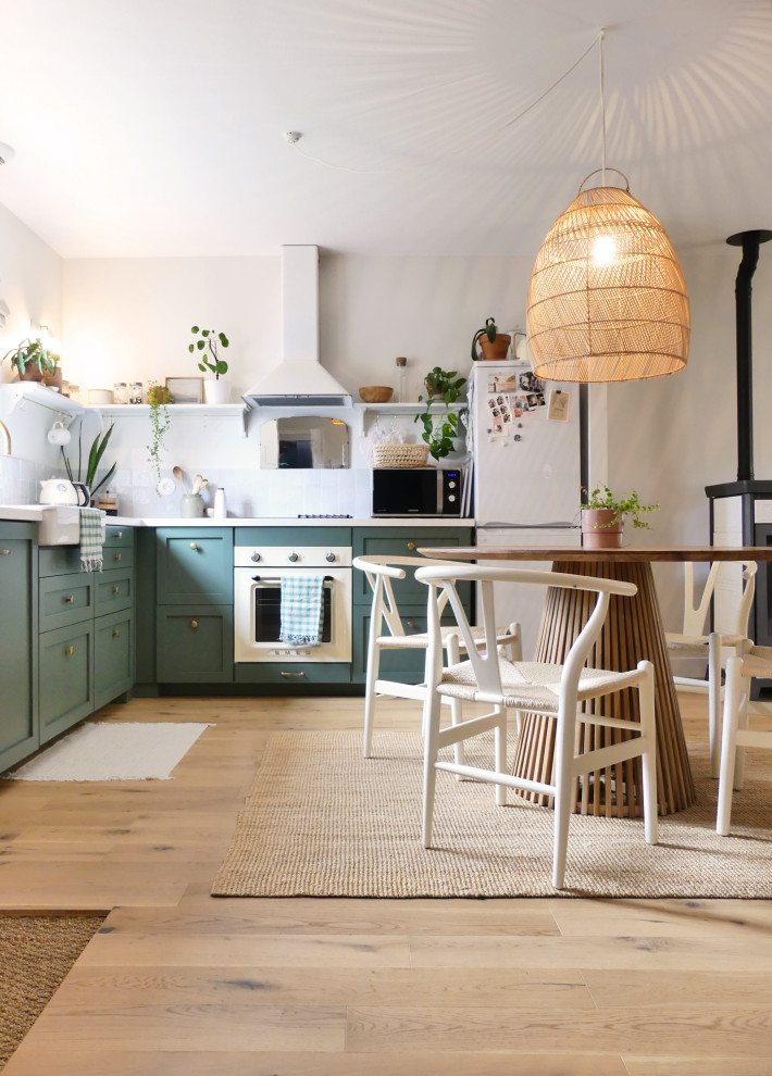 This is an example of a mid-sized beach style l-shaped open plan kitchen in Nantes with a farmhouse sink, green cabinets, laminate benchtops, beige splashback, ceramic splashback, white appliances, light hardwood floors and white benchtop.