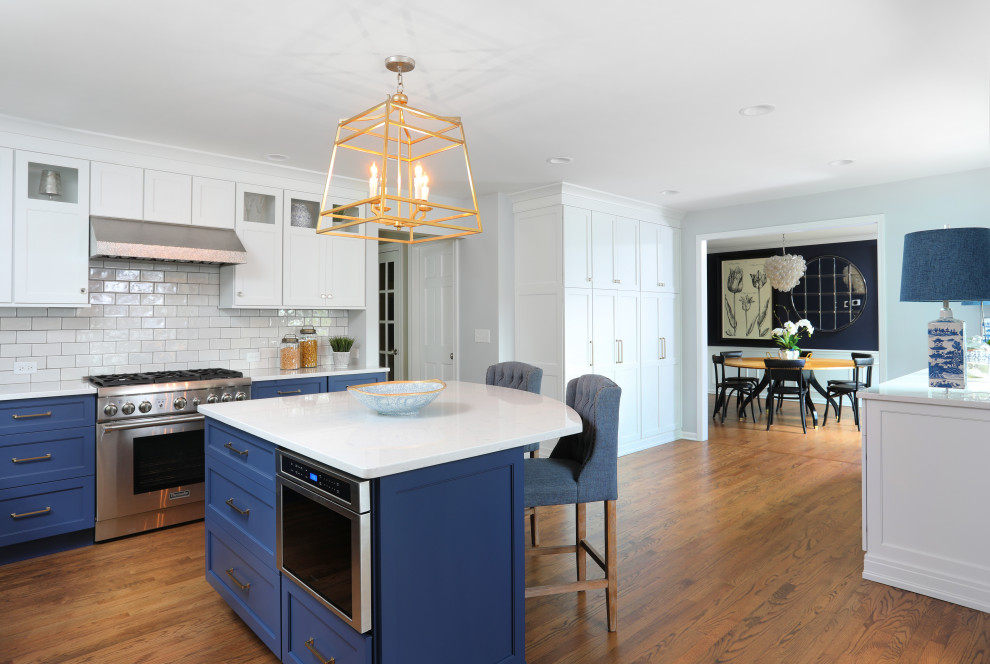 Inspiration for a large transitional l-shaped medium tone wood floor and brown floor eat-in kitchen remodel in Chicago with a farmhouse sink, beaded inset cabinets, blue cabinets, quartz countertops, white backsplash, glass tile backsplash, an island, white countertops and stainless steel appliances
