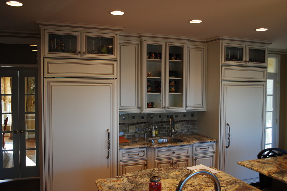 Mid-sized l-shaped medium tone wood floor enclosed kitchen photo with a double-bowl sink, raised-panel cabinets, white cabinets, granite countertops, beige backsplash, stone tile backsplash, stainless steel appliances and an island