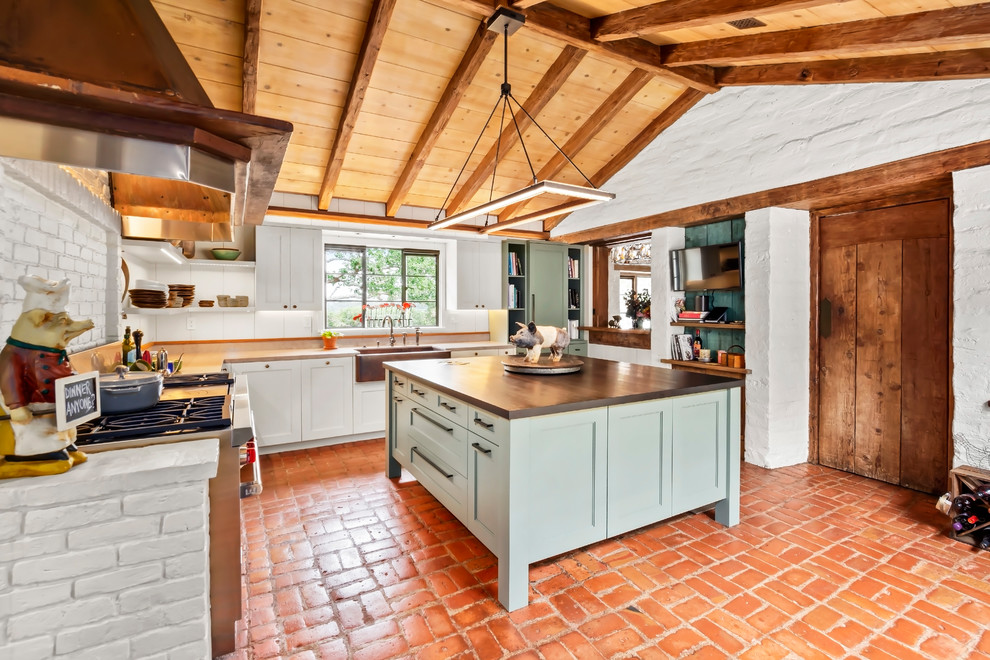 Large arts and crafts u-shaped eat-in kitchen in Other with a farmhouse sink, recessed-panel cabinets, blue cabinets, wood benchtops, stainless steel appliances, brick floors, with island, grey benchtop and white splashback.