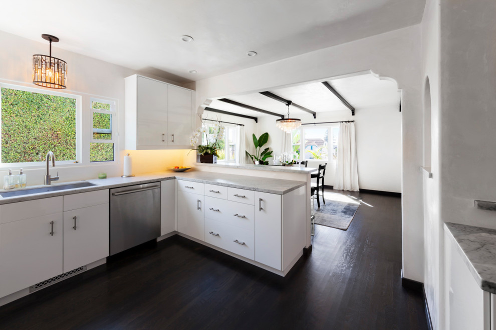 Example of a transitional dark wood floor and brown floor kitchen design in Santa Barbara with white cabinets, marble countertops, stainless steel appliances, a drop-in sink, flat-panel cabinets, white backsplash, a peninsula and white countertops