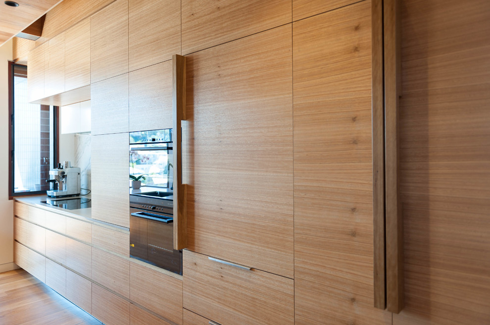 Photo of a mid-sized contemporary galley kitchen pantry in Brisbane with light wood cabinets, quartz benchtops, white splashback, stone slab splashback, black appliances, light hardwood floors and with island.