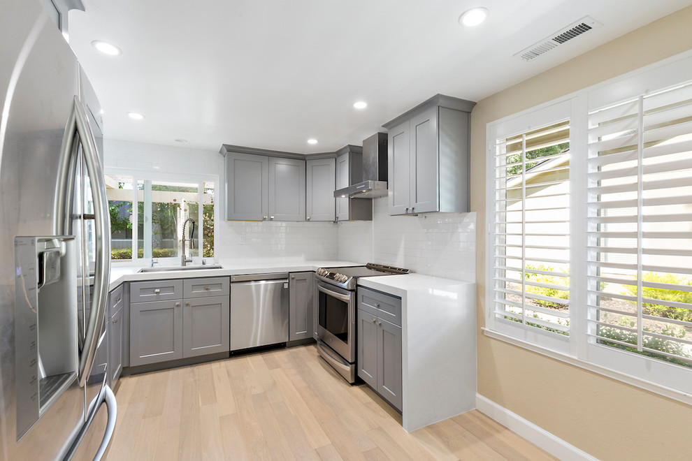 Mid-sized transitional u-shaped light wood floor and beige floor enclosed kitchen photo with an undermount sink, shaker cabinets, gray cabinets, solid surface countertops, white backsplash, stone tile backsplash, stainless steel appliances, no island and white countertops