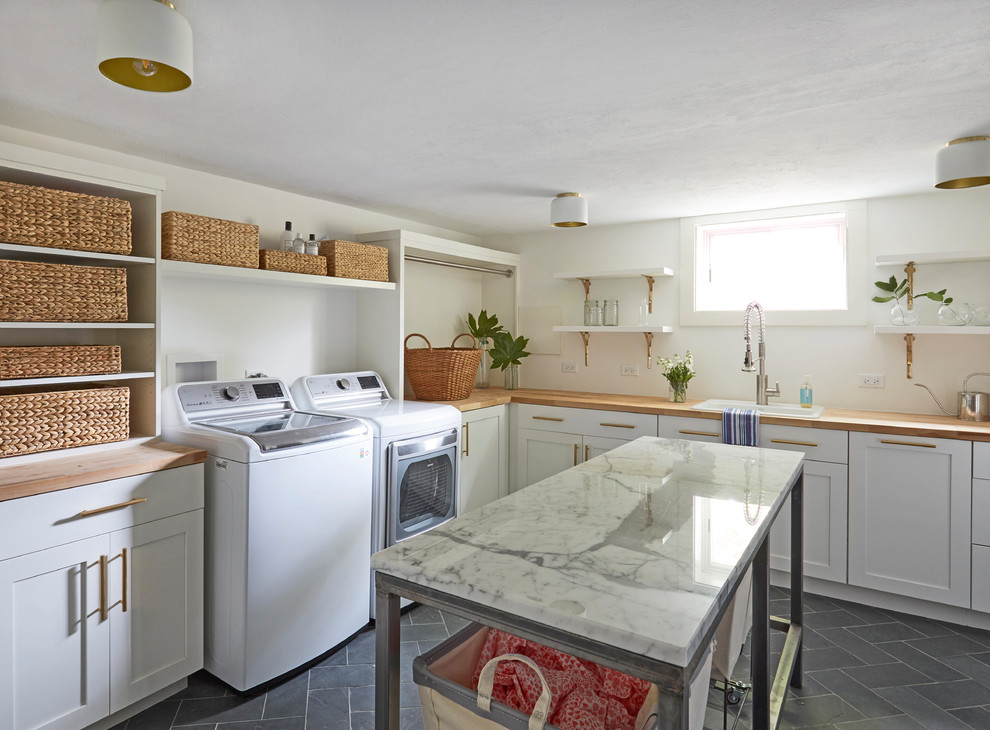 Large transitional l-shaped dedicated laundry room in Chicago with a drop-in sink, shaker cabinets, white cabinets, wood benchtops, white walls, slate floors, a side-by-side washer and dryer, grey floor and brown benchtop.