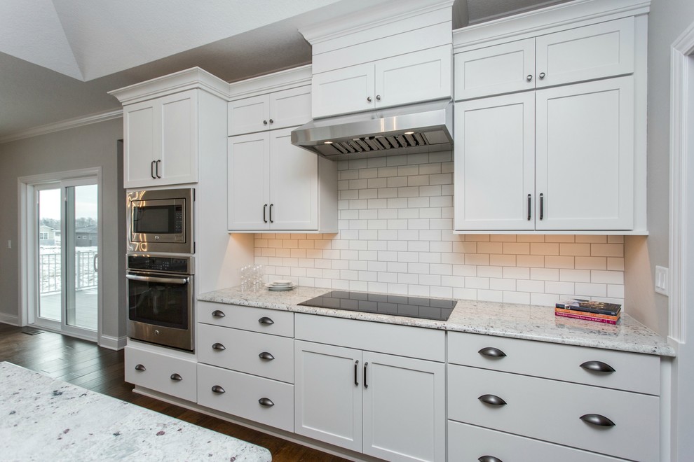 Example of a mid-sized transitional single-wall medium tone wood floor and beige floor open concept kitchen design with a farmhouse sink, recessed-panel cabinets, white cabinets, granite countertops, white backsplash, ceramic backsplash, stainless steel appliances and an island