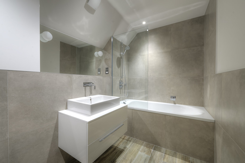 This is an example of a mid-sized contemporary master bathroom in London with a trough sink, a wall-mount toilet, porcelain tile, grey walls, porcelain floors, flat-panel cabinets, white cabinets, a corner tub, a shower/bathtub combo and gray tile.