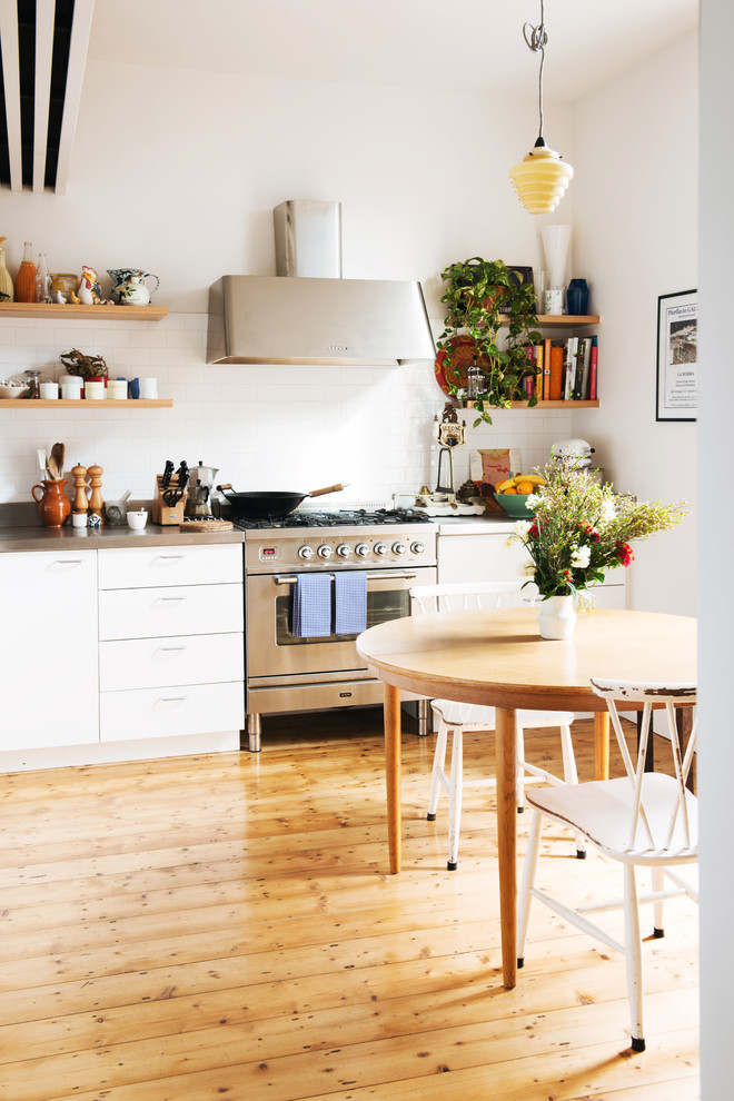 This is an example of a mid-sized scandinavian l-shaped eat-in kitchen in Melbourne with flat-panel cabinets, white cabinets, stainless steel benchtops, white splashback, ceramic splashback, stainless steel appliances, light hardwood floors and no island.