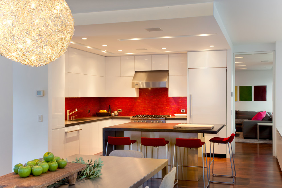 This is an example of a modern l-shaped eat-in kitchen in New York with flat-panel cabinets, white cabinets, concrete benchtops, red splashback and panelled appliances.