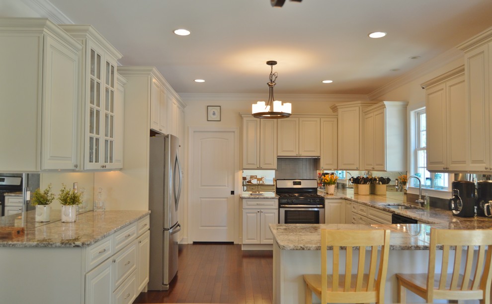 This is an example of a mid-sized arts and crafts galley eat-in kitchen in DC Metro with granite benchtops, stainless steel appliances, raised-panel cabinets, white cabinets, glass sheet splashback, medium hardwood floors, a single-bowl sink, metallic splashback, a peninsula and brown floor.