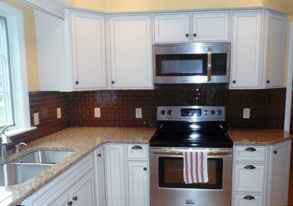 Example of a mid-sized transitional l-shaped laminate floor and yellow floor kitchen pantry design in New York with an undermount sink, recessed-panel cabinets, white cabinets, brown backsplash, ceramic backsplash, stainless steel appliances, no island, beige countertops and granite countertops