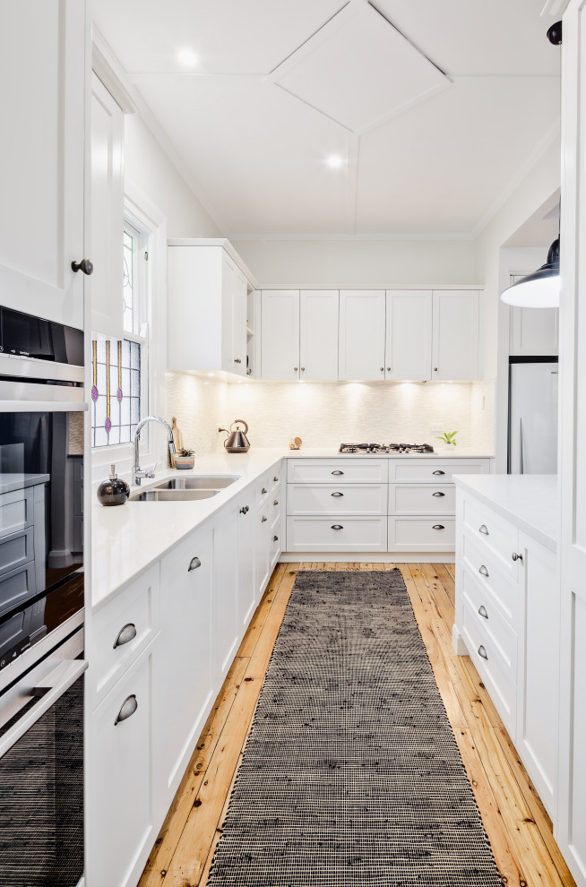 Photo of a mid-sized traditional galley eat-in kitchen in Newcastle - Maitland with an undermount sink, shaker cabinets, white cabinets, quartz benchtops, grey splashback, cement tile splashback, white appliances, light hardwood floors, with island, beige floor and white benchtop.