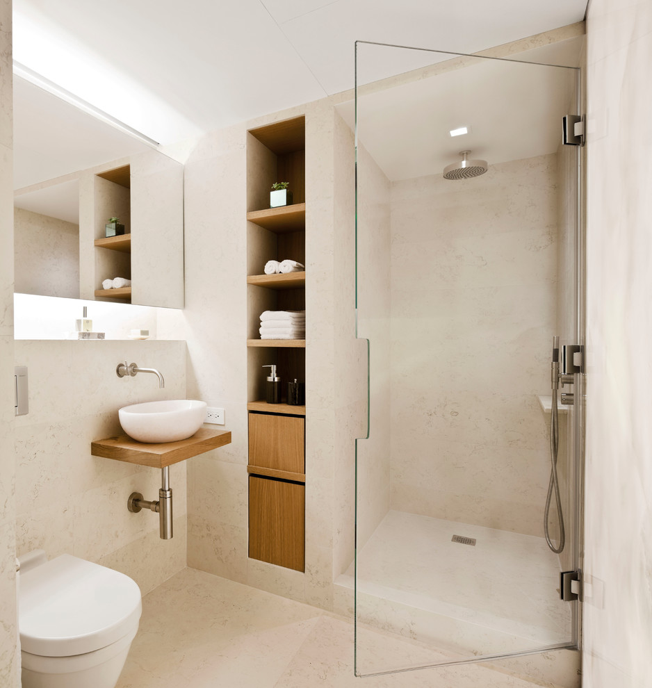 Photo of a mid-sized modern bathroom in New York with a wall-mount toilet, a vessel sink, a hinged shower door, beige walls, an alcove shower, beige tile, limestone, limestone floors, wood benchtops, beige floor and brown benchtops.
