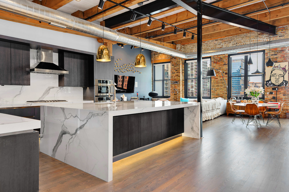 This is an example of an industrial l-shaped eat-in kitchen in Chicago with an undermount sink, flat-panel cabinets, black cabinets, white splashback, stainless steel appliances, light hardwood floors and with island.