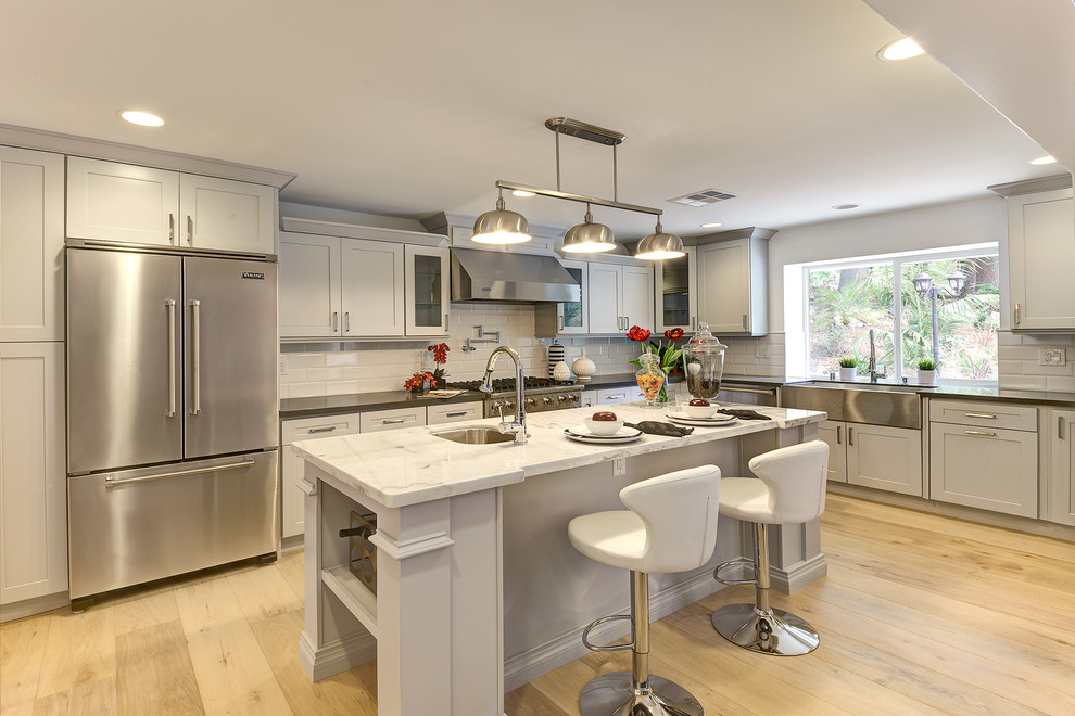Transitional l-shaped kitchen in Los Angeles with a farmhouse sink, shaker cabinets, grey cabinets, white splashback, subway tile splashback, stainless steel appliances, light hardwood floors and with island.