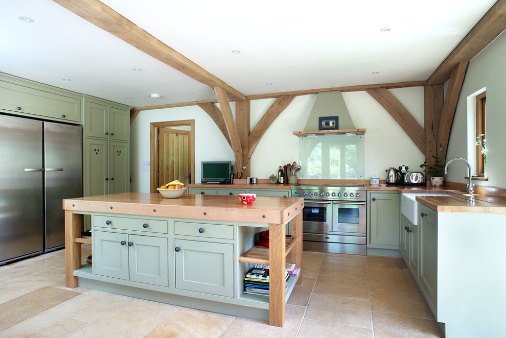 This is an example of a country u-shaped open plan kitchen in Berkshire with limestone floors, a farmhouse sink, beaded inset cabinets, green cabinets, stainless steel appliances, with island and wood benchtops.