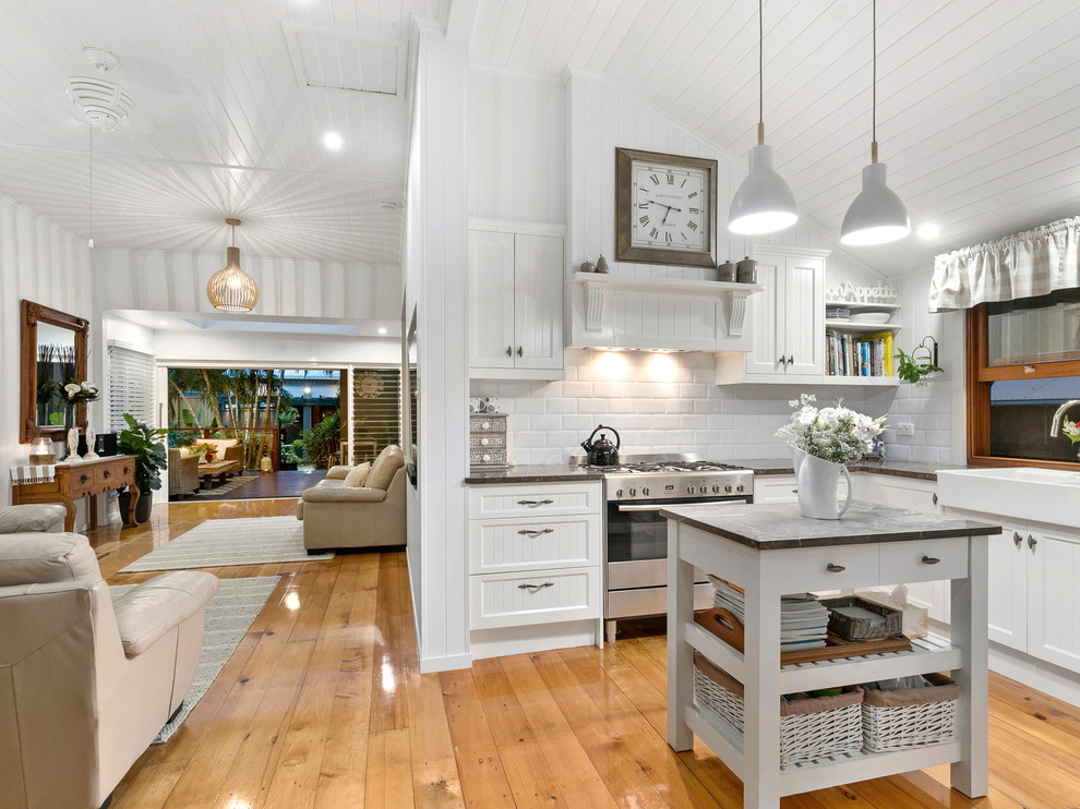 Photo of a small beach style u-shaped open plan kitchen in Brisbane with a farmhouse sink, white cabinets, granite benchtops, white splashback, ceramic splashback, stainless steel appliances, with island, recessed-panel cabinets, medium hardwood floors, brown floor and grey benchtop.