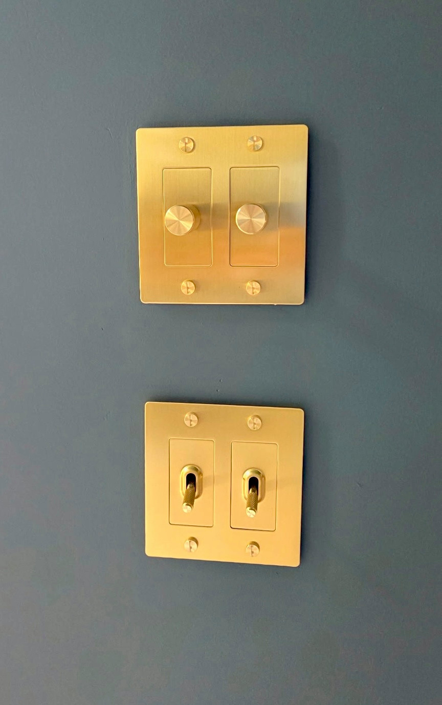 Brushed brass wall hardware, including knurled dimmer switches and toggle switch plates set against dark teal walls and ceilings.