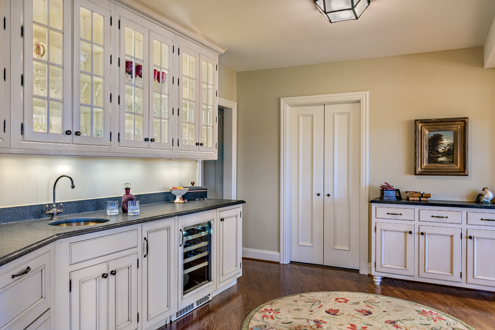 This is an example of a large traditional single-wall wet bar in Nashville with an undermount sink, recessed-panel cabinets, white cabinets, granite benchtops, beige splashback, stone tile splashback, medium hardwood floors, brown floor and black benchtop.