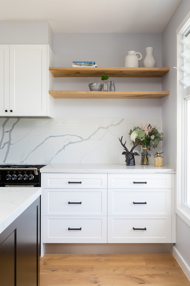 Inspiration for a mid-sized beach style galley open plan kitchen in Gold Coast - Tweed with an integrated sink, raised-panel cabinets, white cabinets, marble benchtops, white splashback, marble splashback, black appliances, medium hardwood floors, with island and brown floor.