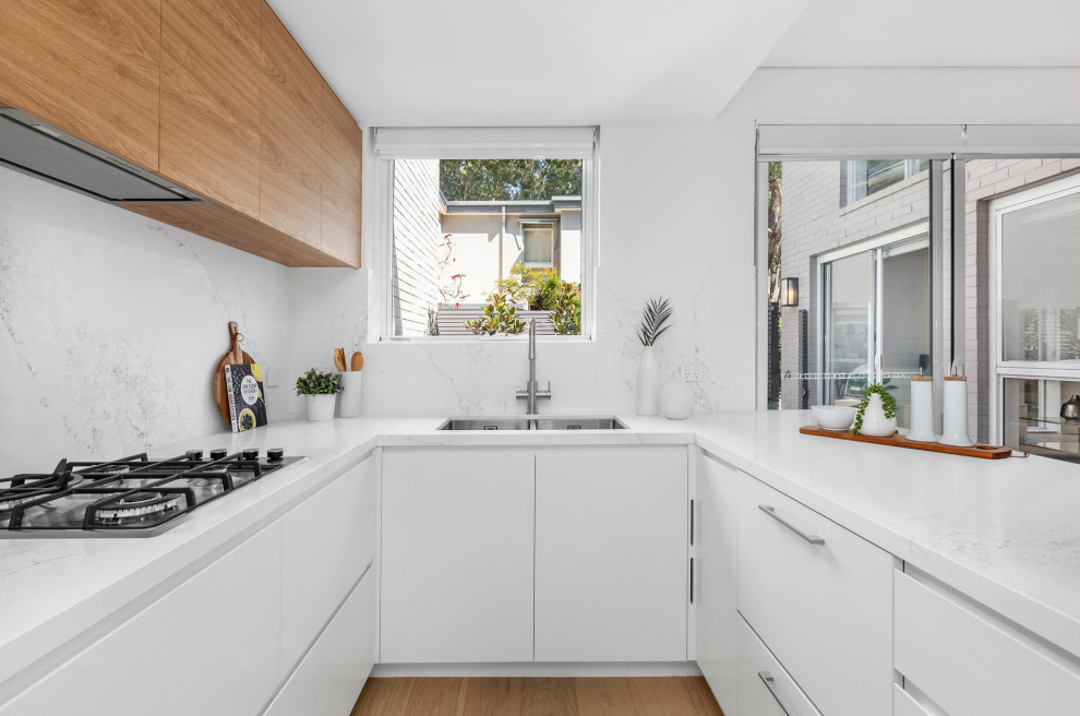 This is an example of a mid-sized modern u-shaped kitchen pantry in Sydney with an undermount sink, flat-panel cabinets, white cabinets, quartz benchtops, white splashback, engineered quartz splashback, stainless steel appliances, laminate floors, a peninsula, brown floor and white benchtop.