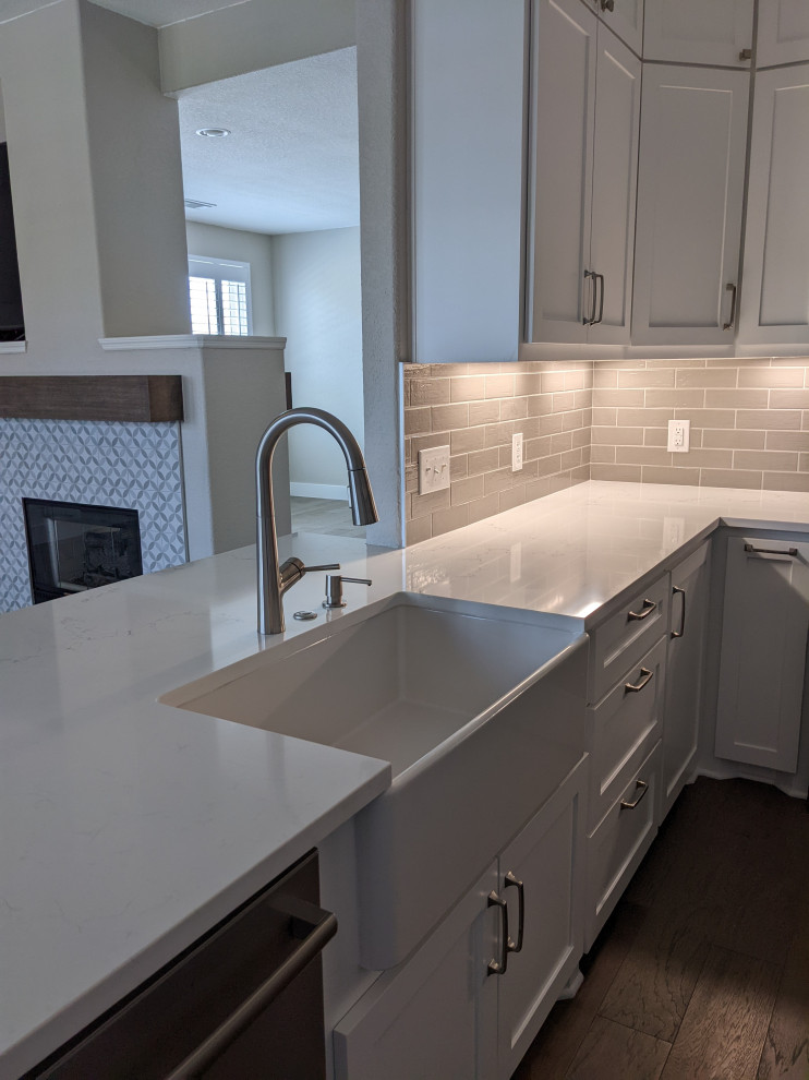 Photo of a mid-sized country u-shaped kitchen in Houston with a farmhouse sink, shaker cabinets, white cabinets, quartz benchtops, grey splashback, subway tile splashback, stainless steel appliances, medium hardwood floors, with island, brown floor and white benchtop.