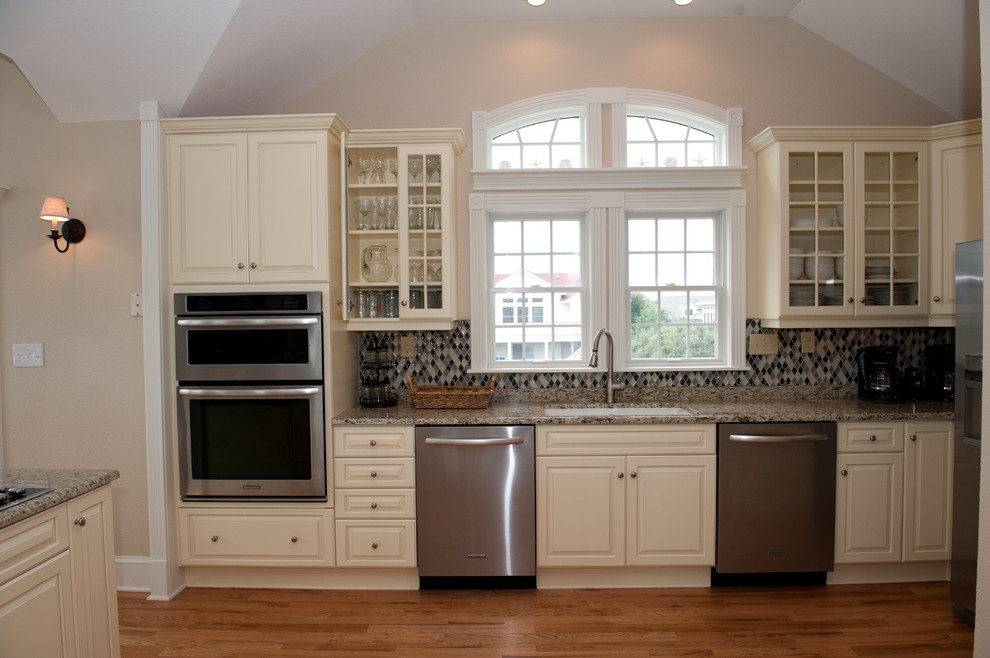 Large transitional light wood floor open concept kitchen photo with an undermount sink, raised-panel cabinets, beige cabinets, granite countertops, multicolored backsplash, mosaic tile backsplash, stainless steel appliances and a peninsula