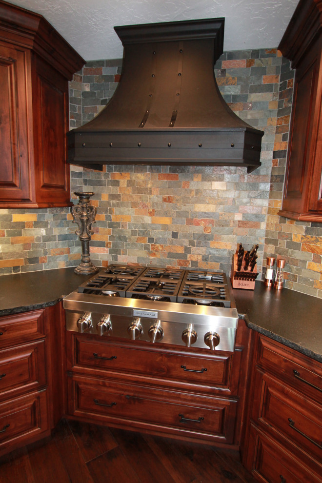 Kitchen - large rustic medium tone wood floor and brown floor kitchen idea with raised-panel cabinets, medium tone wood cabinets, granite countertops, multicolored backsplash, stone tile backsplash, paneled appliances, an island and black countertops