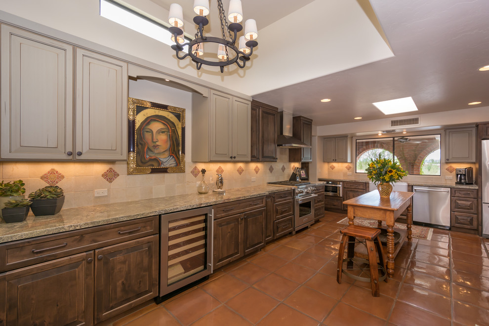 Photo of a mid-sized mediterranean l-shaped separate kitchen in Phoenix with a farmhouse sink, raised-panel cabinets, distressed cabinets, granite benchtops, beige splashback, stone tile splashback, stainless steel appliances, terra-cotta floors, with island and red floor.